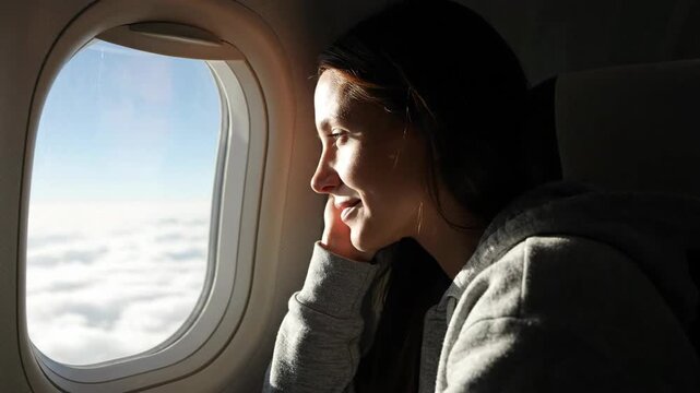 Smiling young woman looking out airplane window at beautiful clouds and blue sky during flight