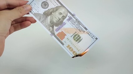 A close-up photo of a US one hundred dollar bill burning in midair against a clean, light background