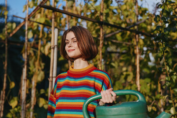 Woman watering plants in garden holding a green watering can, young gardener smiling in a colorful striped sweater among trellis and climbing vines under warm sunlight.