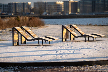Modern wooden empty riverside recreation seats covered in snow standing on riverbank facing bay in middle of city area on sunny day. Public garden near river with sunbeds on low cold winter season.
