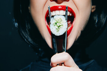 Woman holding sushi with chopsticks, showcasing vibrant red lips and a playful expression on a dark background. Sushi concept in a modern lifestyle setting.