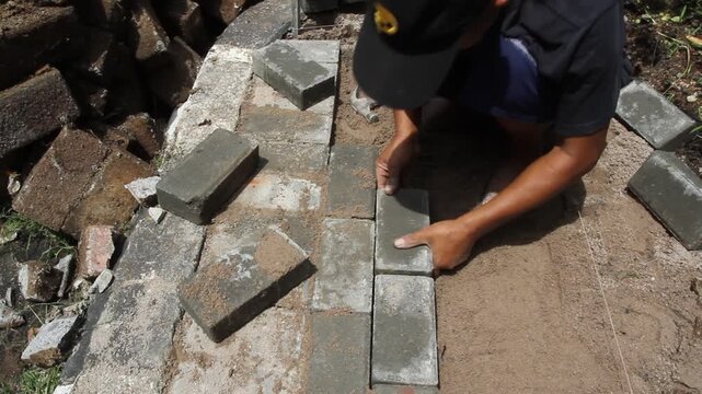 A construction worker interlocking paving stones on a sand bed. A hammer is used to level each paving stone.