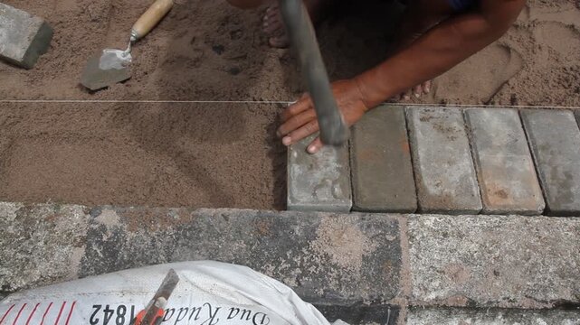 A construction worker interlocking paving stones on a sand bed. A hammer is used to level each paving stone.