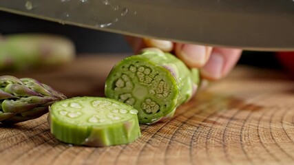 Closeup view of a sharp chefs knife precisely slicing a fresh green vegetable possibly okra or a similar pod on a rustic wooden cutting board highlighting healthy food preparation and culinary skills.
