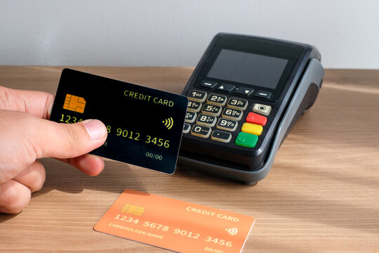 Hand holding yellow and black credit cards in front of an electronic EDC terminal on a wooden desk. Perfect for fintech, payment systems, ecommerce, business finance, banking services, technology - Powered by Adobe