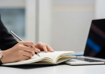 Businesswoman&rsquo;s hands writing notes in a notebook beside a sleek laptop, modern corporate office setting