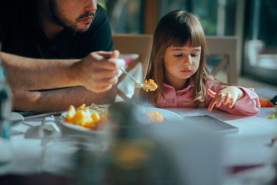 Father Feeding a Kid Who is Playing on the Phone at Mealtime. Little girl being distracted while having lunch engaging with a screen 
