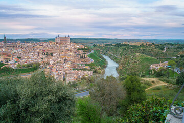 The ancient city of Toledo Spain glows softly at spring twilight with the Tagus River curving around the historic walls forming a natural barrier while lush greenery frames this elevated view.