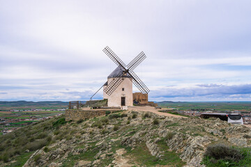 Traditional white windmill with restored sails perched on a rocky hill along the famous Don Quixote Route, overlooking green agricultural plains in spring.