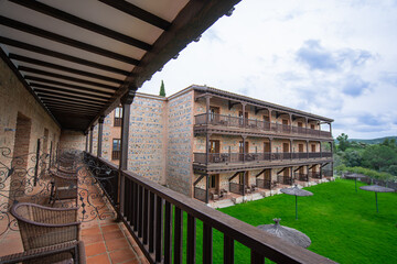 Toledo, Spain - March 13, 2025: Wooden balcony with wicker furniture overlooking three-story stone castle hotel with arcade balconies and lawn.