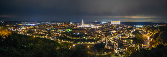 Panoramic night view of Toledo from Mirador del Valle in spring. The illuminated historic city with Alcazar and cathedral glows above the Tagus River bend under dramatic twilight sky.