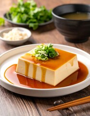 A close-up shot of a square, golden-hued food item drizzled with glossy sauce and topped with green onions. Served on a white plate with accompanying ingredients