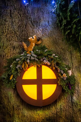Whimsical squirrel decoration above glowing round window on tree trunk with pine branches and lights creating cozy fairytale christmas mood at night