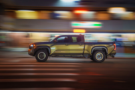 LOS ANGELES, CA - December 25, 2025: Green Toyota Tacoma pickup truck driving through colorful street intersection at night. Blurred motion