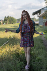 Young woman with long red hair wearing a floral dress and denim jacket walks along abandoned railway tracks surrounded by greenery, capturing a moment of carefree exploration