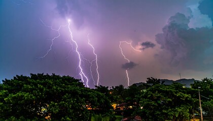 A night scene captures multiple lightning strikes illuminating a dark sky above lush green trees and hinting at a distant landscape