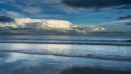 Fototapeta premium Early morning. Dawn over the ocean. The waves roll towards the shore, spreading across the beach. The clouds in the blue sky are highlighted in gold. Reflection on wet, smooth sand. Malaysia. Borneo.