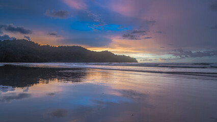 Beautiful tropical sunset over the ocean. Pastel shades. Waves are spreading across the sandy beach. The clouds in the blue sky are highlighted in pink. Reflection on wet, smooth sand. Long exposure. 