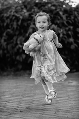 Young girl with a teddy bear running joyfully across a grassy field, wearing a light dress and sneakers, capturing the essence of childhood playfulness and freedom