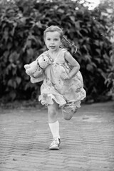 Young girl with a teddy bear running joyfully across a grassy field, wearing a light dress and sneakers, capturing the essence of childhood playfulness and freedom