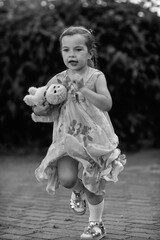 Young girl with a teddy bear running joyfully across a grassy field, wearing a light dress and sneakers, capturing the essence of childhood playfulness and freedom