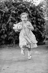 Young girl with a teddy bear running joyfully across a grassy field, wearing a light dress and sneakers, capturing the essence of childhood playfulness and freedom