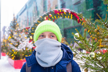 Portrait of a smiling teenager wearing a scarf covering half his face in a city decorated for Christmas on a snowy winter day.