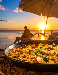 A close-up paella dish on a woven mat, with a blurred beach scene. A person sits enjoying the sunset under an umbrella