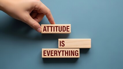 A hand placing wooden blocks spelling "ATTITUDE IS EVERYTHING" on a blue background, conveying a motivational message.