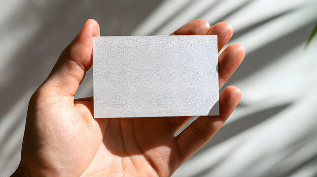 Person holding a blank textured white business card mockup displaying subtle roughness under dramatic sunlight with tropical leaf shadows for branding presentation - Powered by Adobe