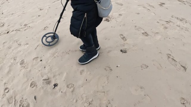 Person walking across soft beach sand while sweeping a metal detector over scattered footprints, searching for lost items during a calm winter outing.