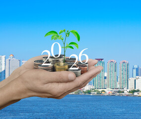 Hands holding growing tree with 2026 white text and coins over city tower, skyscraper and river, Happy new year 2026 business ecological concept