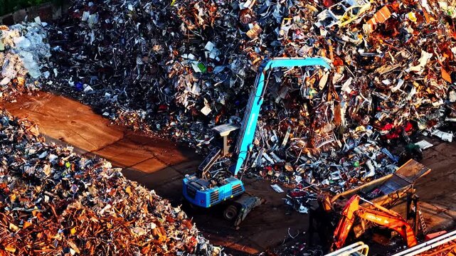 Aerial view of a blue material handler sorting a massive heap of mixed scrap metal at an industrial recycling yard, surrounded by piles of crushed debris and machinery.