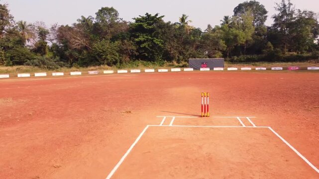 Cinematic drone shot of a red soil cricket pitch with yellow wickets in a rural tropical setting. Perfect for sports, travel, and cultural content.