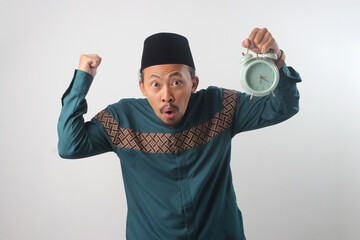 Excited Asian Muslim man in green traditional outfit holding an alarm clock. Isolated on white...