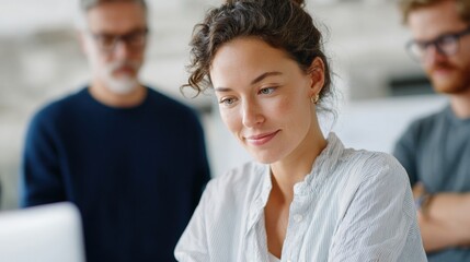 Young woman with curly hair, wearing a white blouse, sitting in front of a laptop computer. she is looking off to the side with a slight smile on her face.