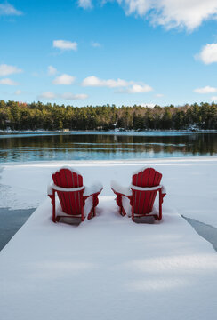 Snow covered red chairs on end of dock facing icy lake in winter.