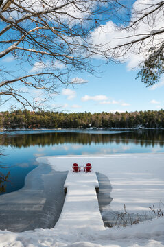 Snow covered red chairs on end of dock facing icy lake in winter.