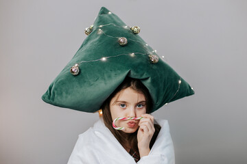 Child wearing Christmas tree pillow hat and holding candy cane indoors