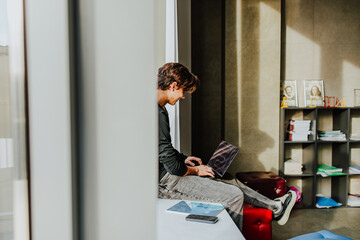 Student sitting by window in classroom using laptop and smiling