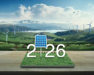 2026 white text with solar cell on green grass on open book on wooden table over panoramic view of wind turbine farm on mountain, Happy new year 2026 green ecology and saving energy concept