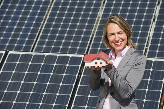 Person holding miniature house model in front of solar panels outdoors