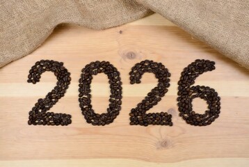 The numerals "2026" creatively formed by arranging roasted coffee beans on a wooden table, set against a softly blurred cozy caf&eacute; interior background with warm lighting. A few lighter beans add subtle