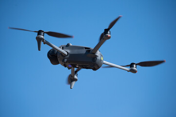 Bottom view of a modern grey quadcopter drone flying against a clear blue sky. Professional unmanned aerial vehicle with four propellers for video and photography.