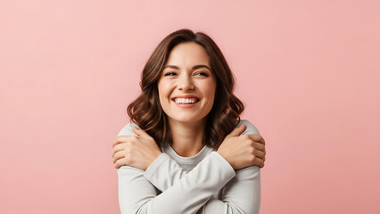 Fototapeta premium A beautiful woman with long brown hair smiling genuinely while embracing herself warmly against a pink backdrop.