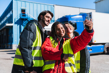 Diverse group of smiling truck drivers and logistics workers taking a selfie, wearing safety vests, celebrating teamwork at facility