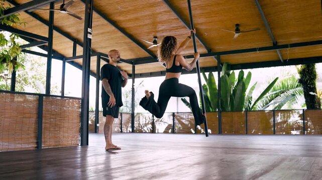 Determined athletic woman performing climbing exercises on a pole during a private workout session with her male personal trainer in a spacious tropical studio