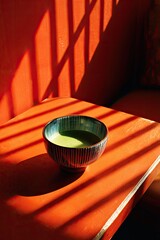 A bowl of green tea sits on an orange surface, shadowed, red wall