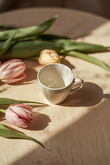 A delicate white coffee cup adorned with intricate handle details and a subtle sheen, filled with a warm brown liquid, sits elegantly beside a bouquet of fresh tulips in various shades of pink 