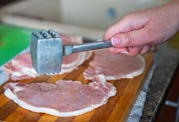 A woman tenderizes fresh raw pork chops with a metal mallet on a wooden cutting board. The process of cooking a homemade meal in the kitchen. The process of cooking schnitzel. 